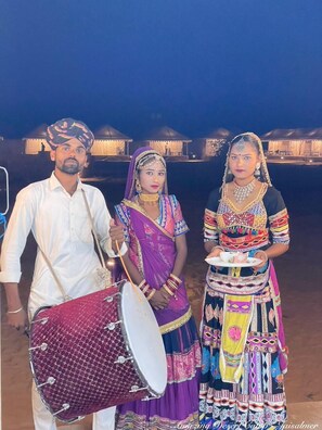 A group of people in traditional attire, one playing a drum, standing in an outdoor setting with tents in the background. - Amazing Desert Camp (Jaisalmer)