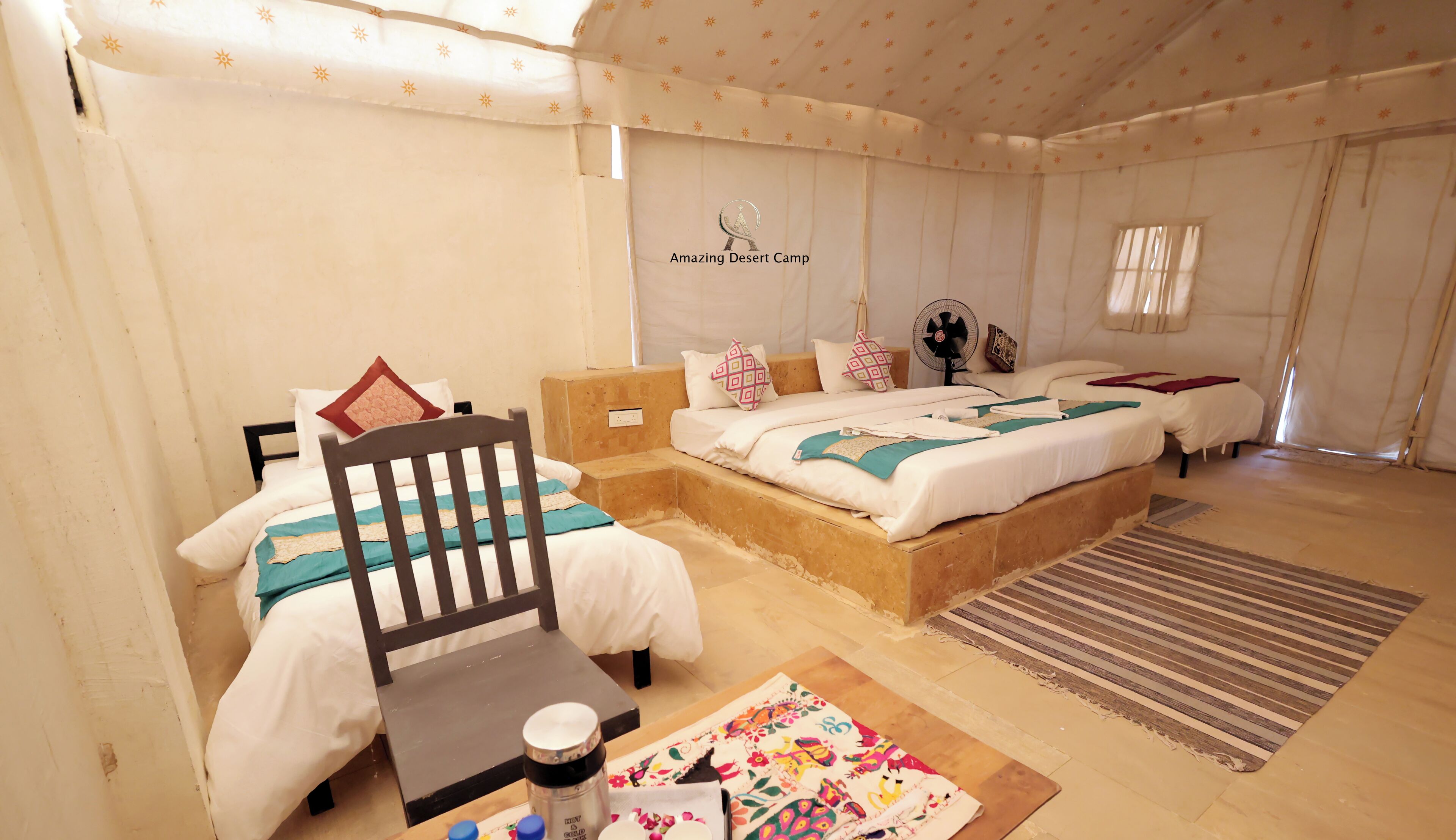 A desert camp room with two beds, a wooden chair, a fan, and a table with a bottle and a cloth.