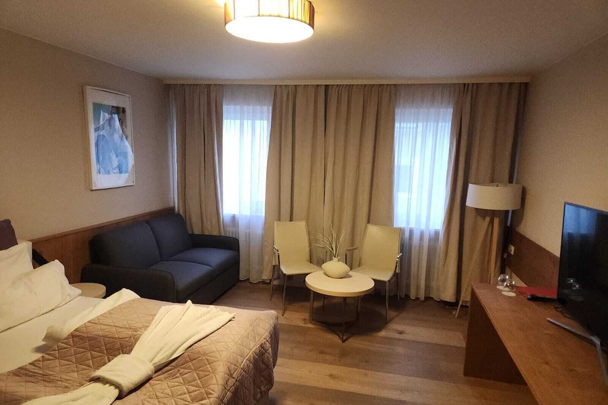 A hotel room with a bed, a sofa, two chairs, a small table, a TV, and curtains.