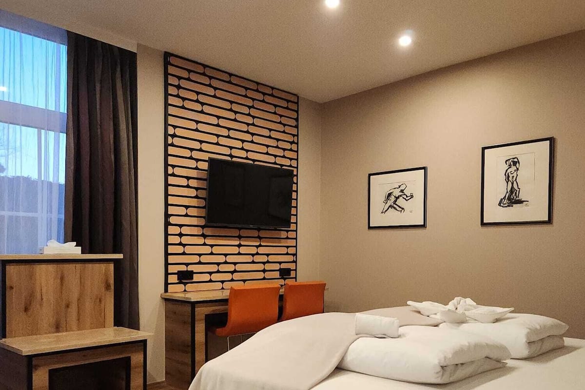 A modern hotel room with a large bed, a desk with a chair, a flat-screen TV mounted on a brick-patterned wall, and two framed artworks on the wall.