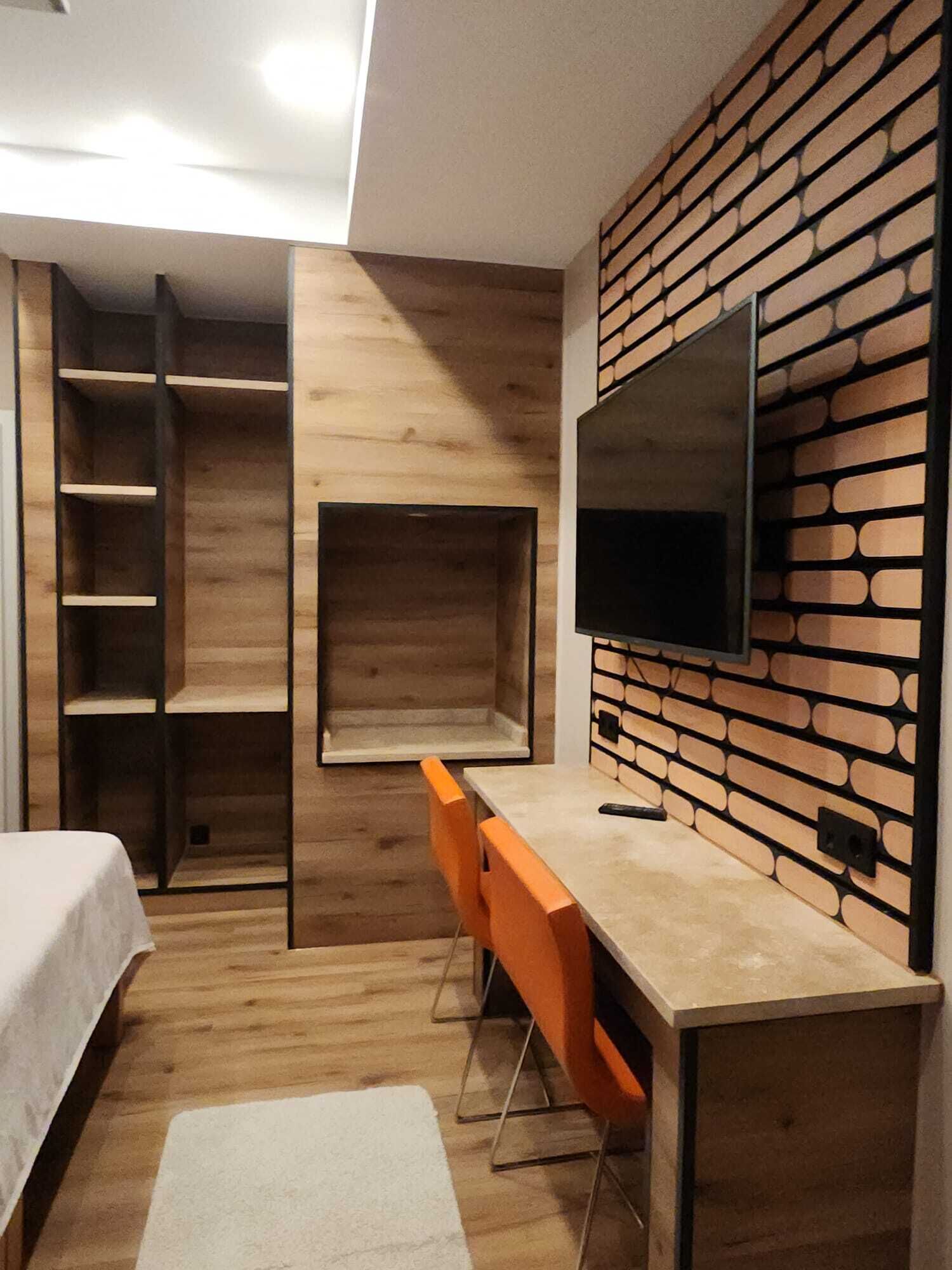 A modern room with a bed, a desk with a chair, a TV mounted on a brick-patterned wall, and a built-in shelving unit.