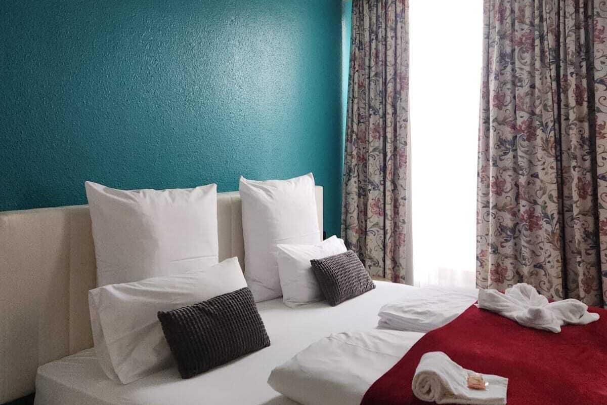 A hotel room with a teal accent wall, a bed with white linens, a red throw blanket, and floral curtains.