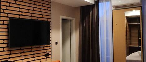 Habitación de hotel con un televisor de pantalla plana montado en una pared con diseño de ladrillo, una cama con cubrecama blanco, dos sillas naranjas y un escritorio de madera.