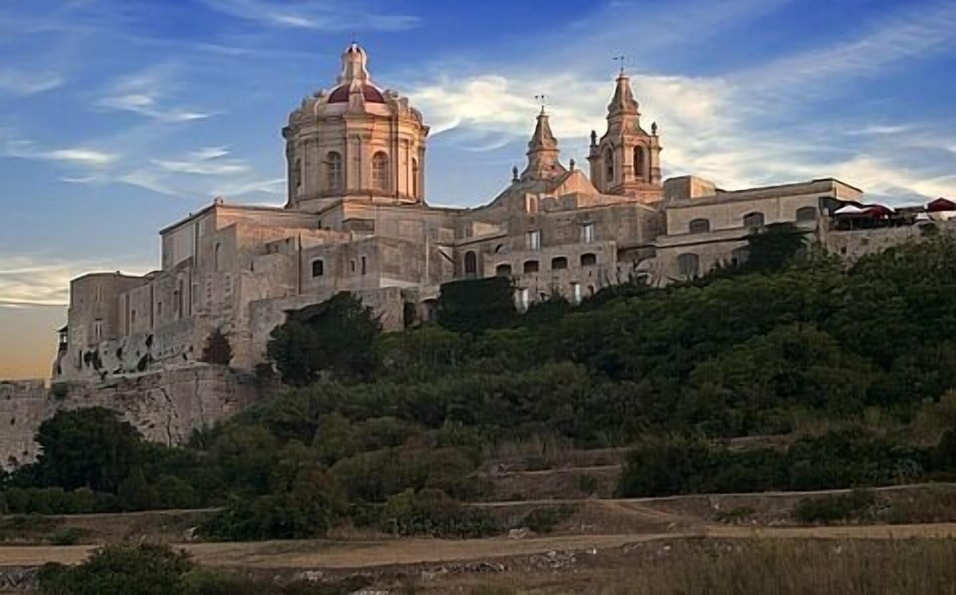 A historic building with multiple domes and towers, situated on a hill with surrounding greenery.