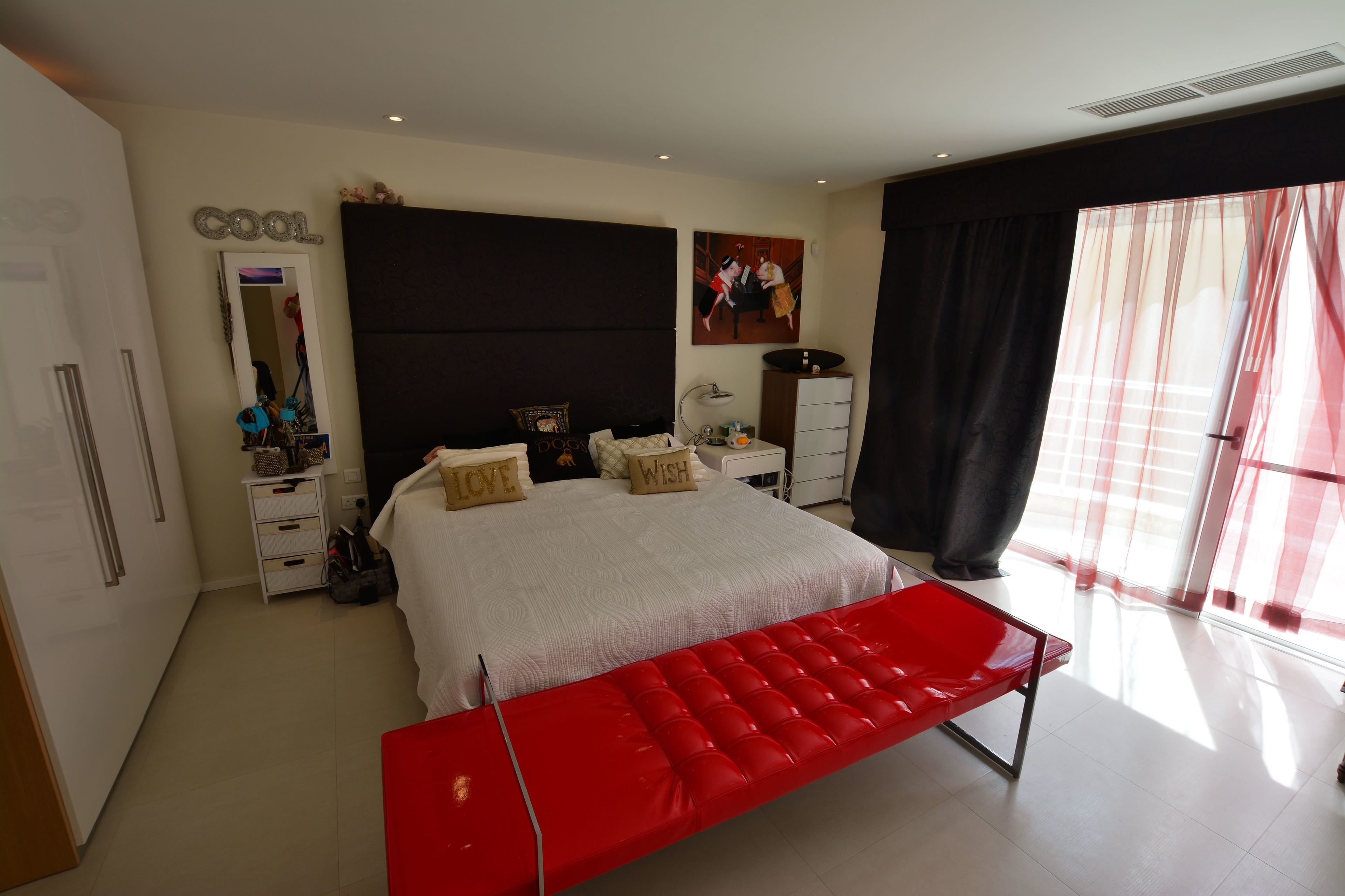Un dormitorio con una cama grande, un banco rojo, un armario, un espejo, una cómoda y un cuadro en la pared.