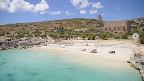 A beach with clear turquoise water, a few people sunbathing, and a building with a tower in the background. - Super 3 Bedroom Villa With Amazing Ocean Views (Swieqi)