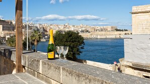 A bottle of Cinzano Prosecco wine and a glass on a stone ledge overlooking a city with historic buildings and a body of water. - Traditional Maltese Townhouse, Roof Terrace and Views (Senglea)