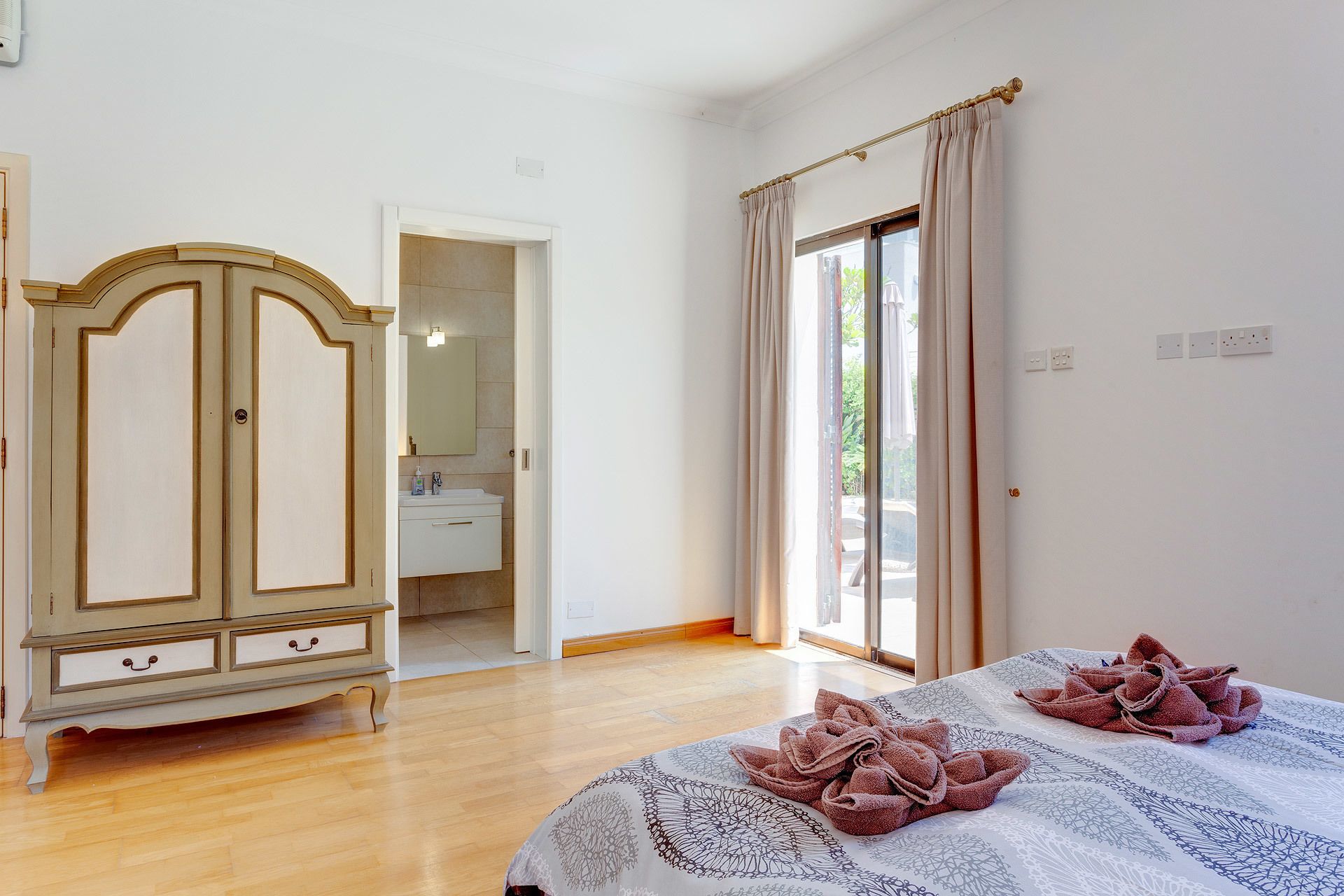 Une chambre à coucher avec un lit, une armoire, une salle de bain visible à travers une porte ouverte et une fenêtre avec des rideaux.