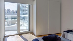 A modern bedroom with a large window, a bed with a blue and gray bedspread, a white ceiling light, and a view of the cityscape. - Modern Apartment With Stunning Seaviews (Sliema)
