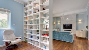 Un salon avec une bibliothèque blanche, un canapé bleu, un fauteuil blanc et un téléviseur fixé sur un mur en briques blanches.