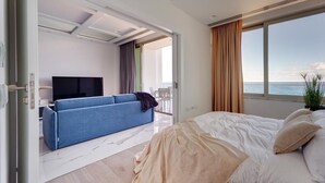 A modern hotel room with a bed, a sofa, a TV, and a balcony with a view of the sea.
