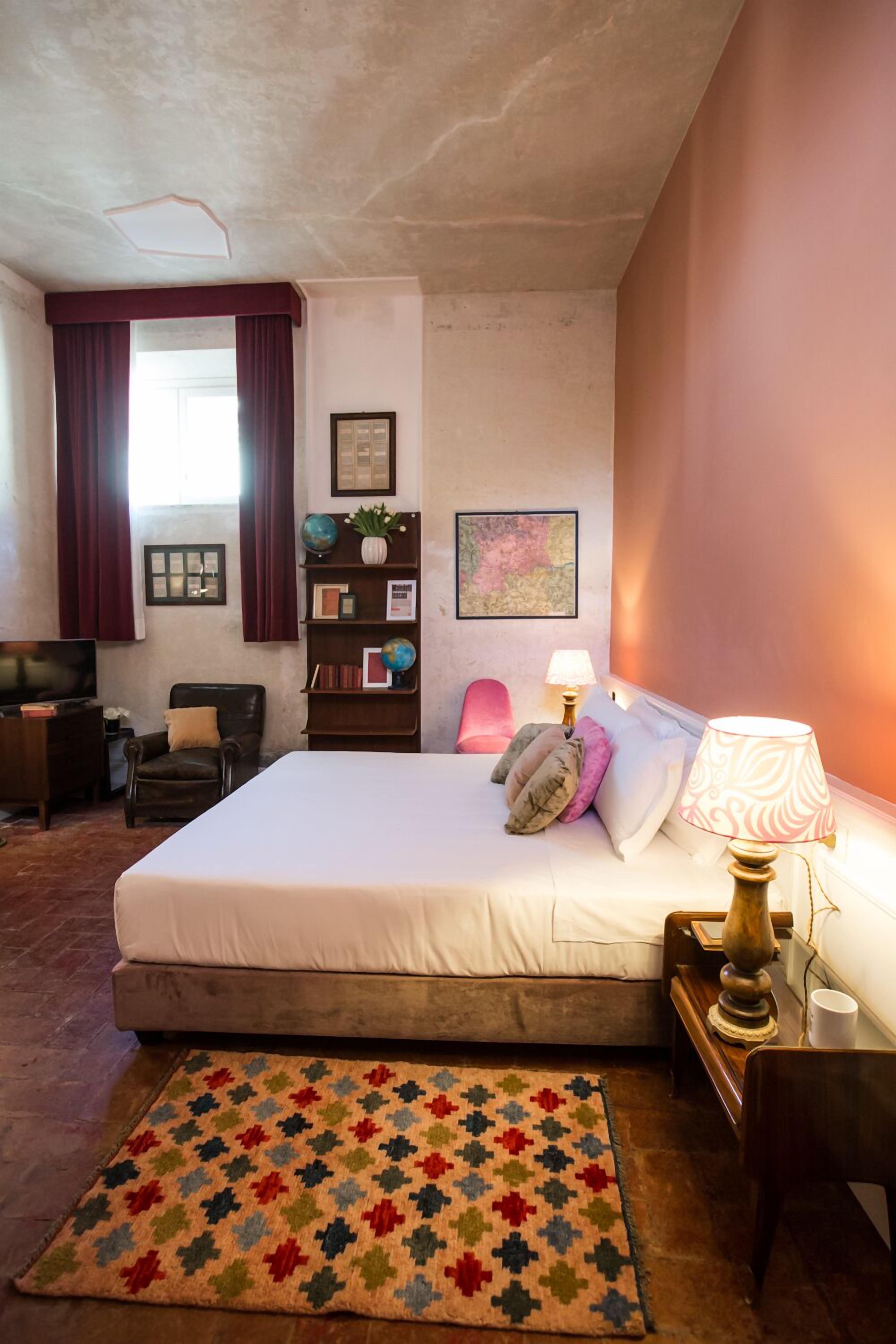 Un dormitorio con cama, escritorio, silla, televisor, estantería y ventana con cortinas.