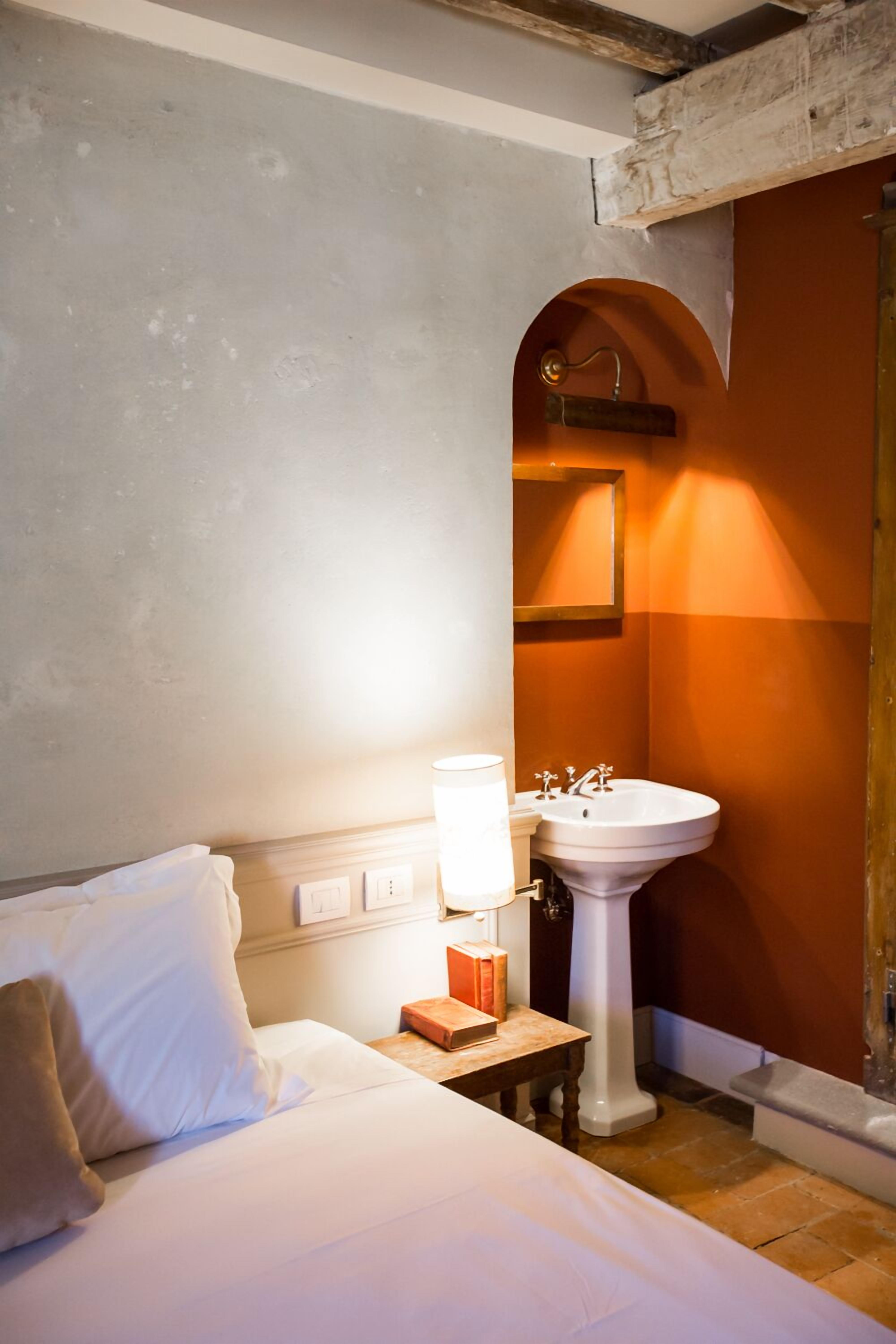 Un dormitorio con cama, mesita de noche, lámpara y un lavabo blanco con espejo.