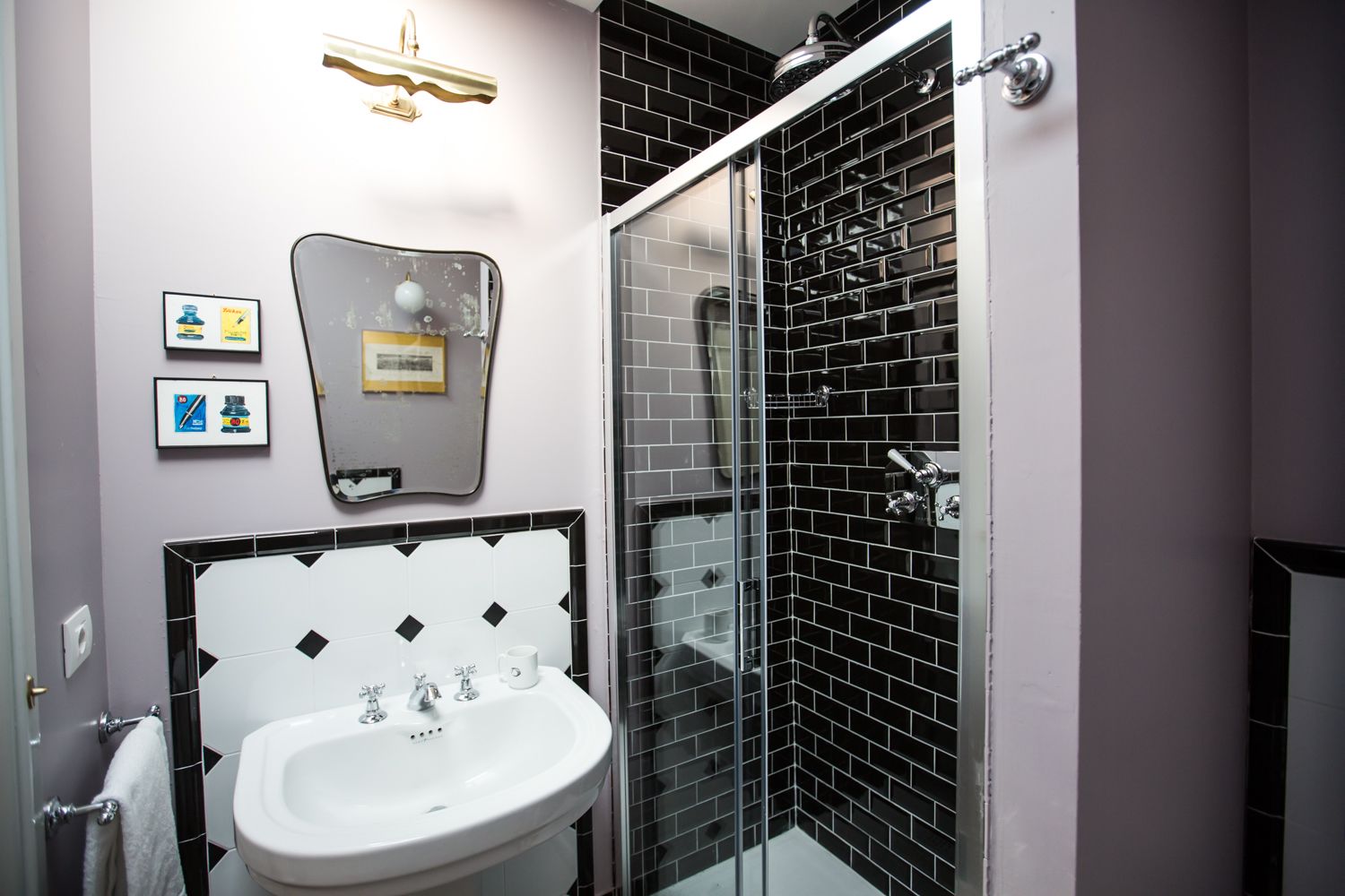 Une salle de bain avec un lavabo blanc, un miroir doté d’un cadre décoratif, des murs carrelés en noir et blanc, et un espace douche avec des carreaux noirs.