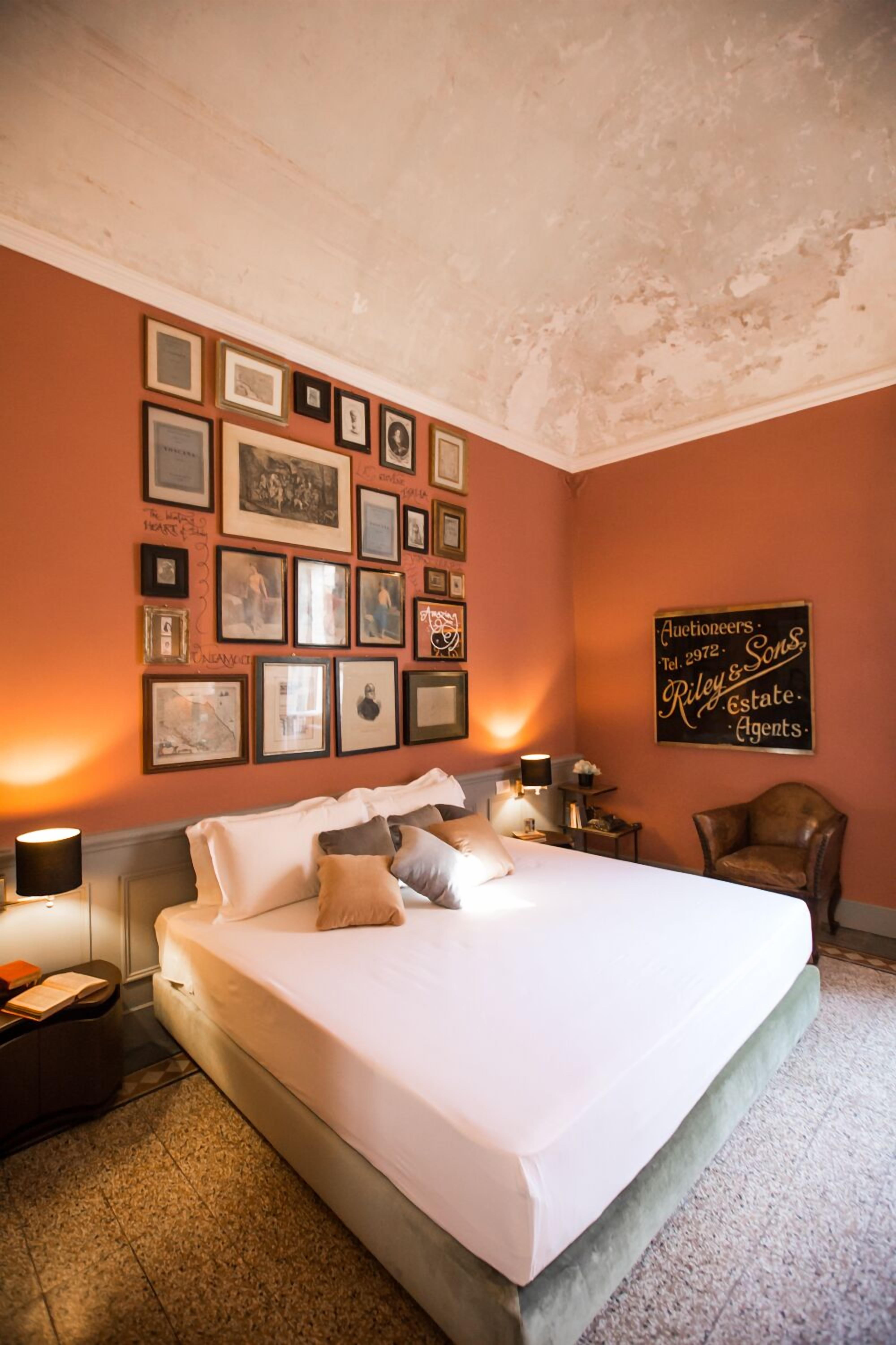 Une chambre d’hôtel avec un grand lit, des tableaux encadrés au mur, une enseigne indiquant « Riley & Sons, commissaires-priseurs », et un fauteuil en cuir.