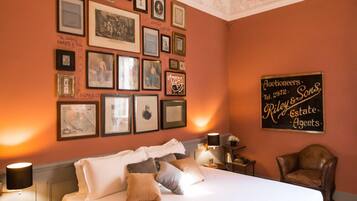 Une chambre d’hôtel avec un grand lit, des tableaux encadrés au mur, une enseigne indiquant « Riley & Sons, commissaires-priseurs », et un fauteuil en cuir.