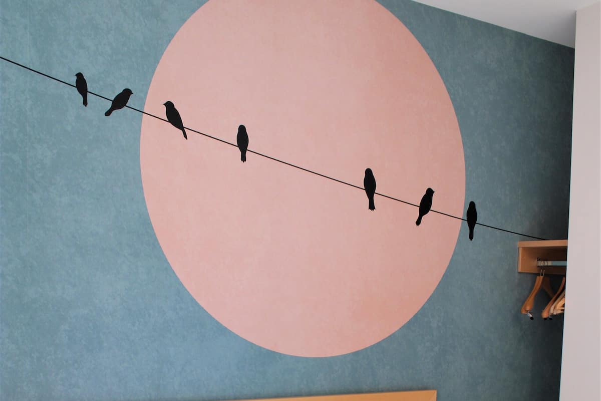 Une chambre à coucher avec une tête de lit en bois, une grande fresque murale représentant des oiseaux perchés sur un fil électrique, et un lit recouvert de draps blancs.