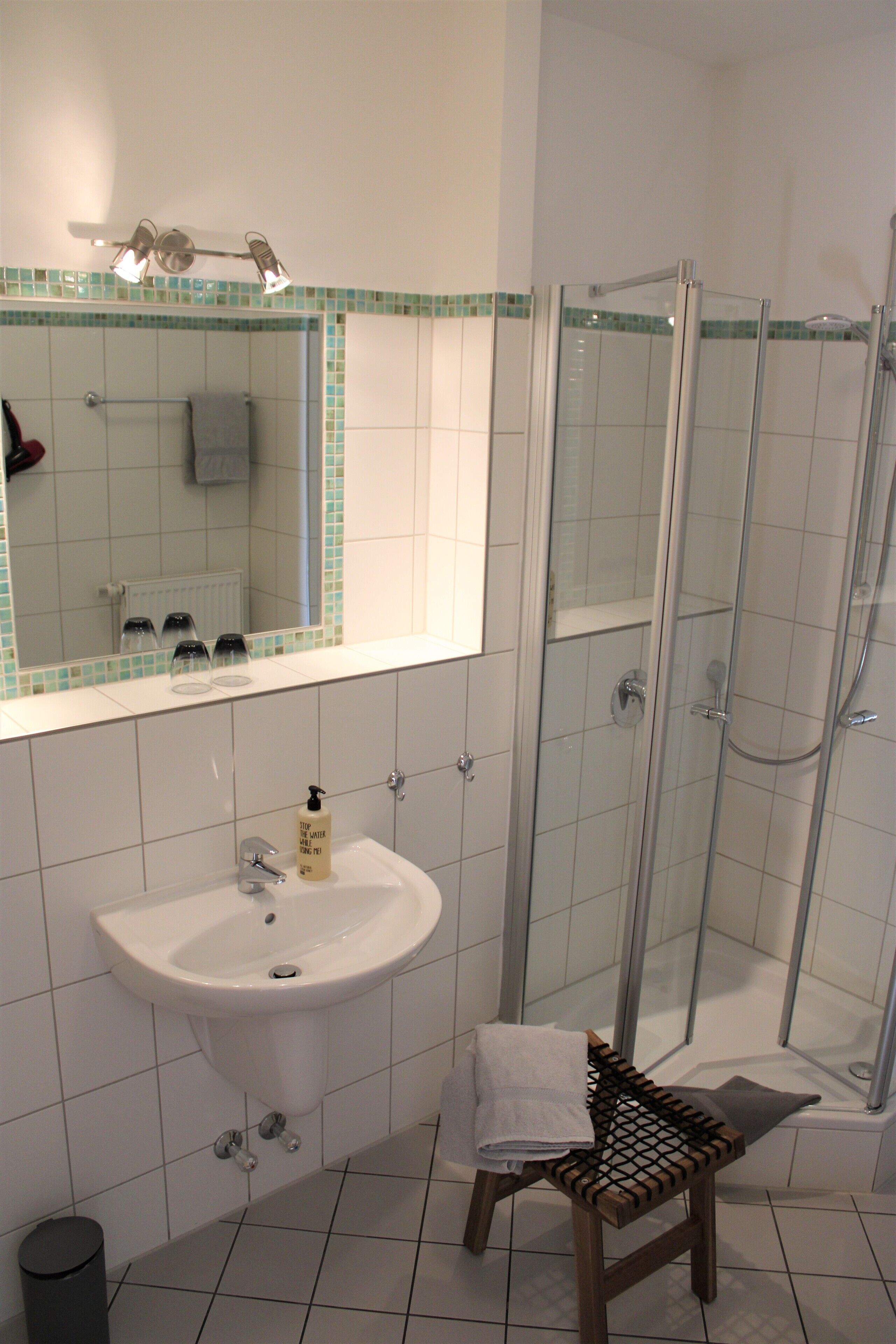 Un baño con lavamanos blanco, un espejo, cabina de ducha y un taburete pequeño.