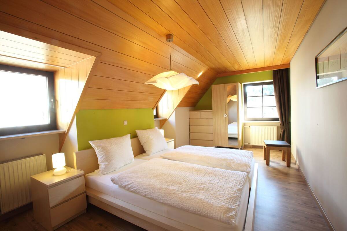 Une chambre avec un plafond en pente, un lit recouvert de linge de lit blanc, une table de chevet avec une lampe et une fenêtre avec des rideaux.