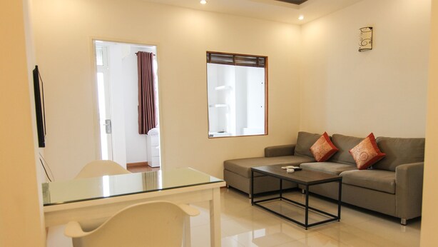 A modern living room with a glass dining table, a white chair, a grey sofa with red cushions, and a large window with blinds.