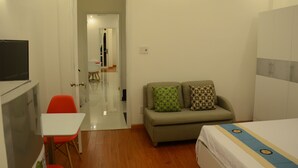 A small, well-lit room with a bed, a sofa with patterned cushions, a red chair, and a television on a stand.