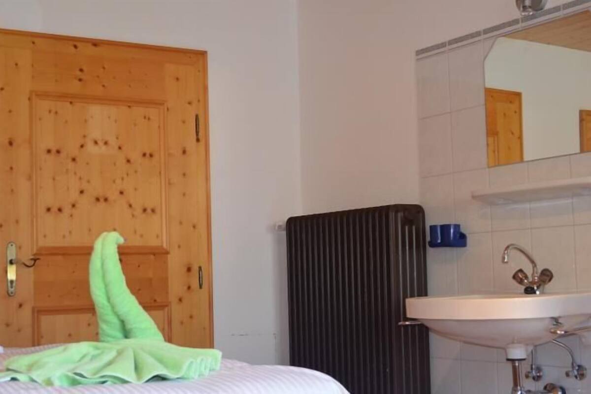 A bedroom with a wooden door, a radiator, a bed with a green blanket, and a bathroom with a sink and mirror.