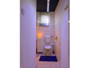 Ein schmales Badezimmer mit Toilette, einem kleinen Schrank und einem Fenster mit Jalousien.