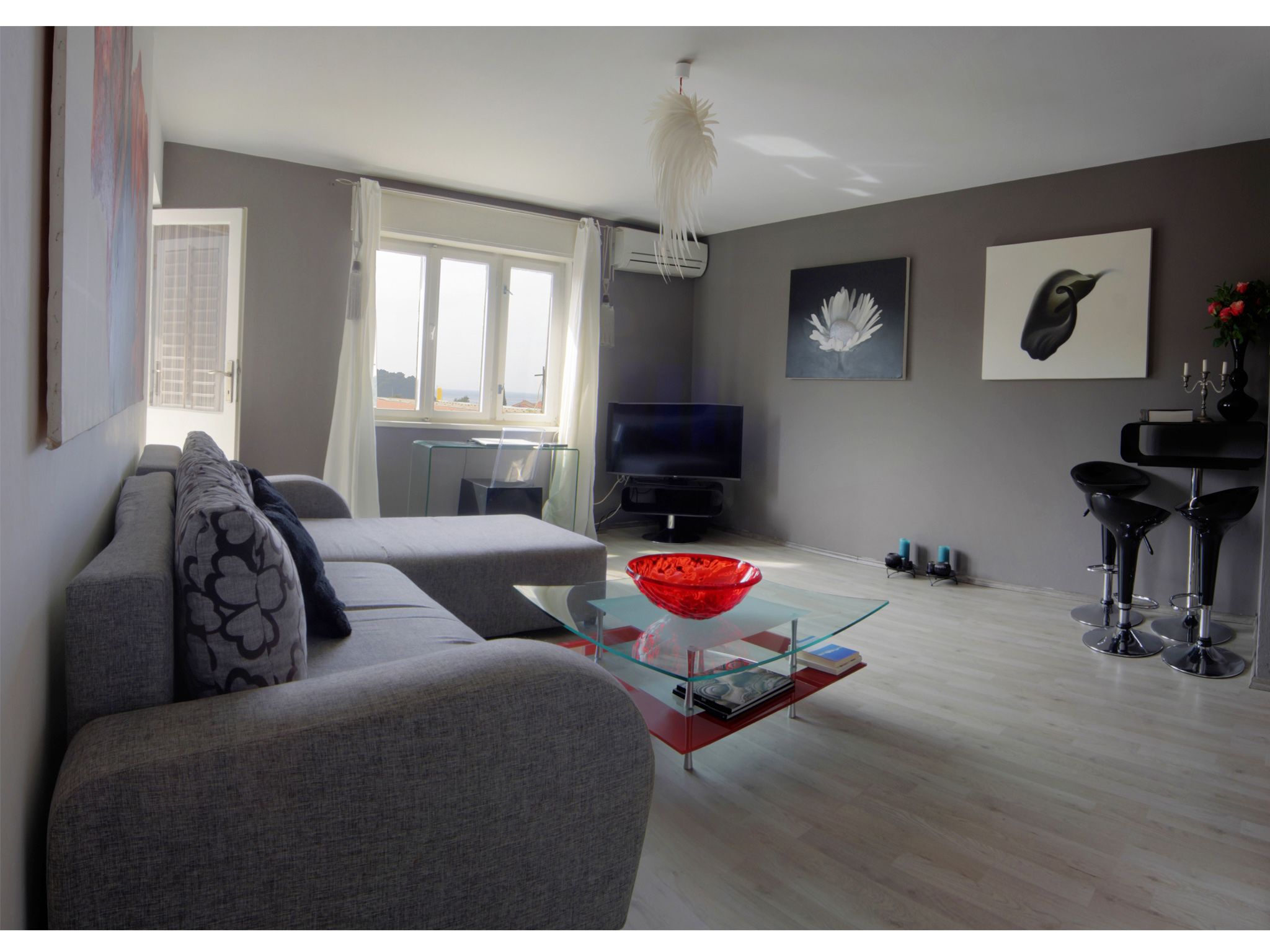 A modern living room with a grey sofa, a glass coffee table with a red bowl, a flat-screen TV, and a dining area with black bar stools.