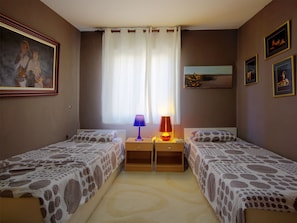 Ein Schlafzimmer mit zwei Betten, einem Nachttisch mit Lampe, einem Fenster mit Vorhängen und gerahmten Bildern an den Wänden.
