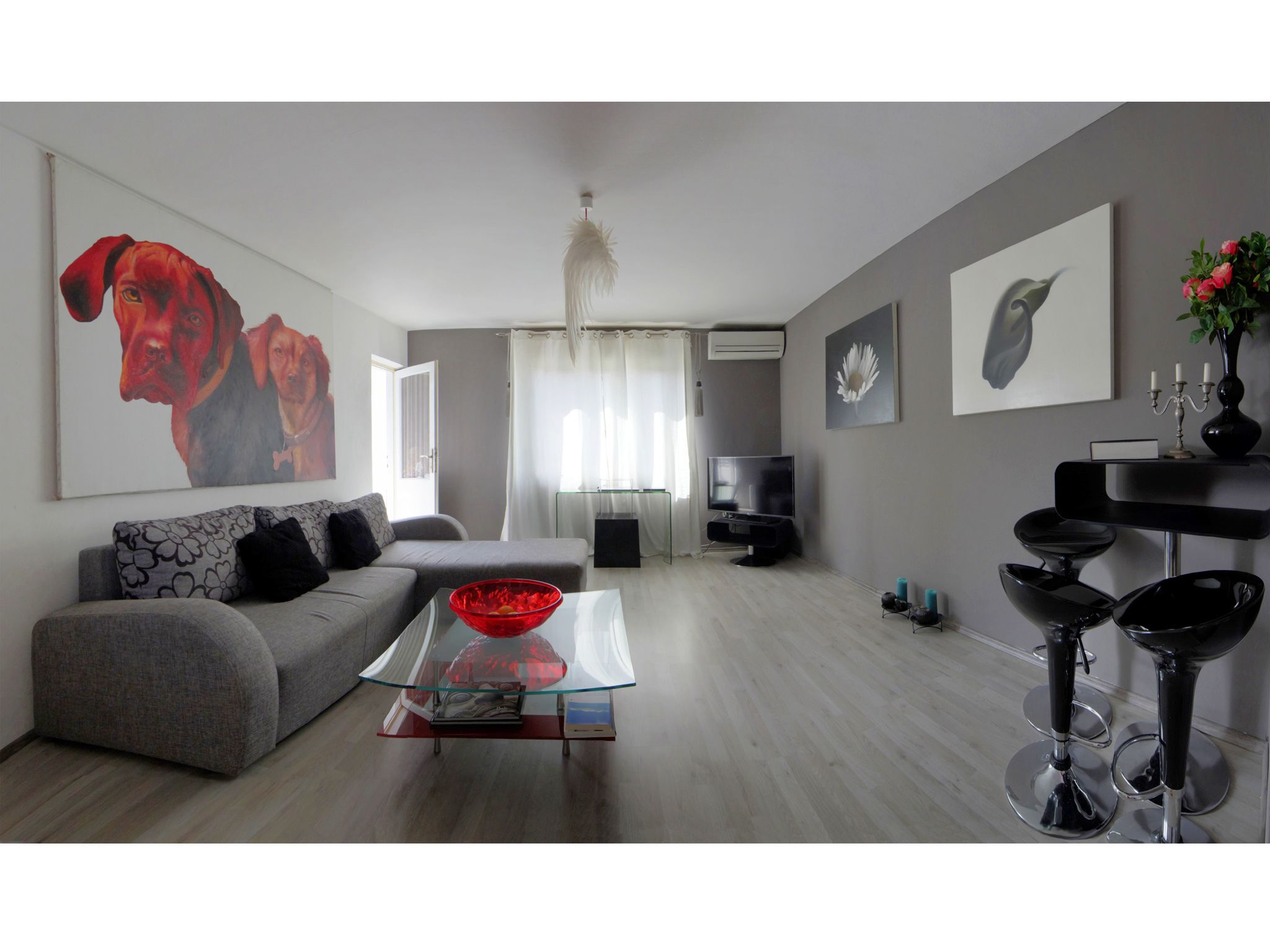 A modern living room with a grey sofa, a glass coffee table, a TV, and a dining area with bar stools.