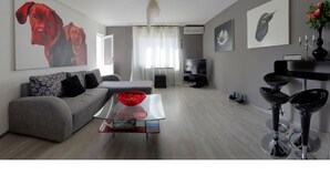 A modern living room with a grey sofa, a glass coffee table, a TV, and a dining area with bar stools. - Art Apartment (Rovinj)