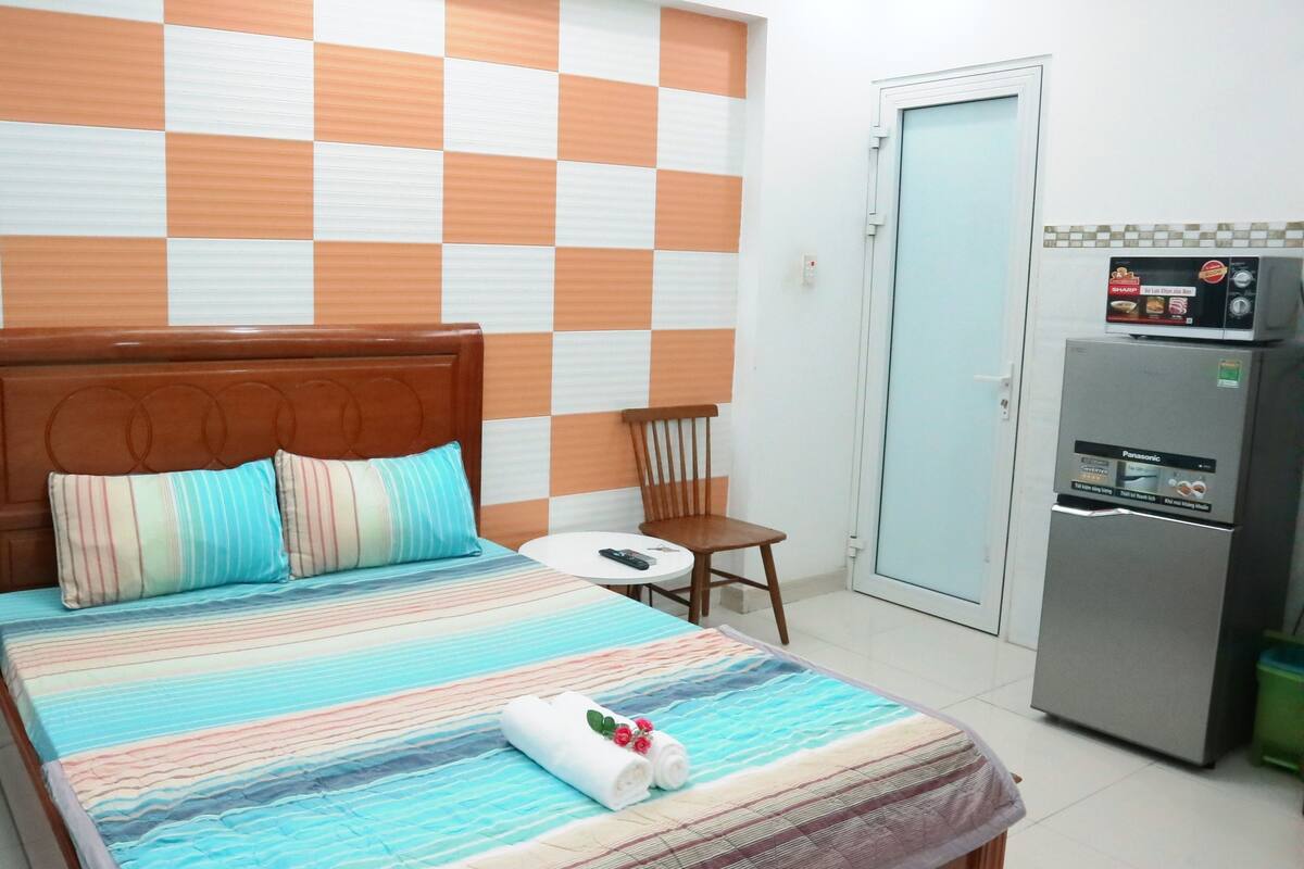 A hotel room with a checkered wall pattern, a bed with striped bedding, a wooden headboard, a bedside table with a remote control, a chair, a refrigerator, and a fan.