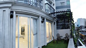 A modern building with large sliding glass doors leading to a well-lit interior, a small outdoor seating area with chairs, and a green artificial turf lawn. - Smiley Apartment 8 (Ho Chi Minh City)