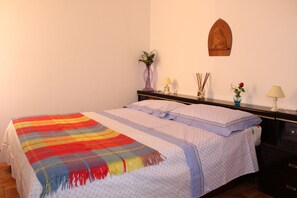 Una cama bien hecha con un edredón colorido, una mesita de noche con un florero y un relieve de madera en la pared.
