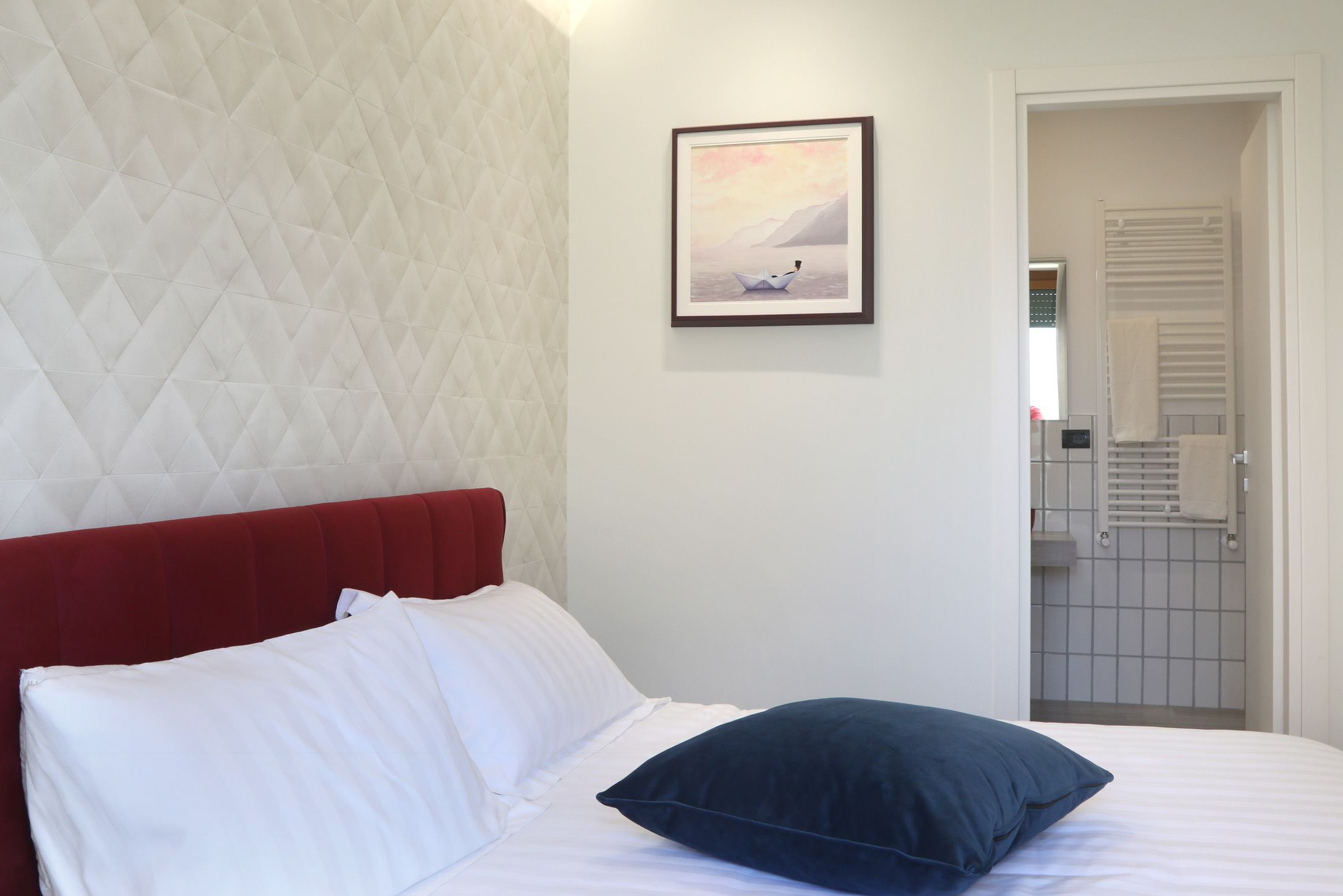 Ein Hotelzimmer mit rotem Kopfteil, weißer Bettwäsche, einem gerahmten Bild an der Wand und Blick ins Badezimmer durch eine geöffnete Tür.