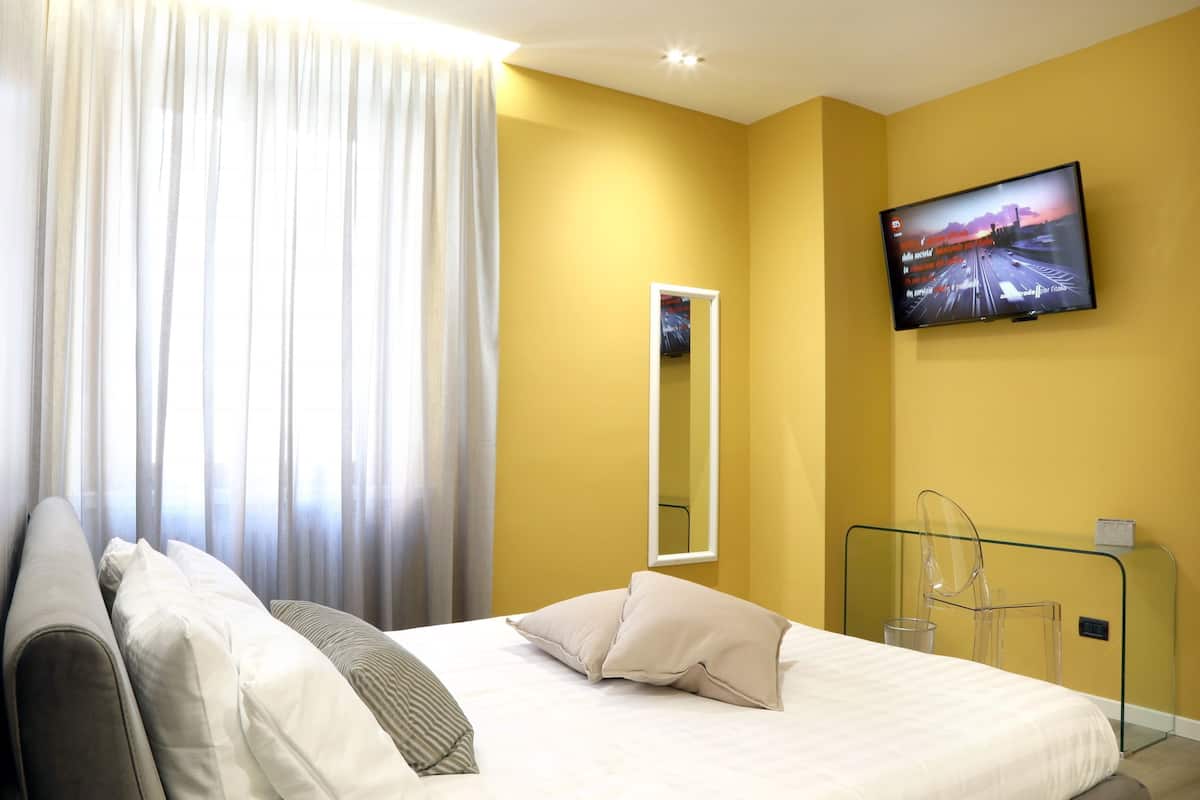 A hotel room with a bed, a television mounted on the wall, a mirror, and a glass partition.
