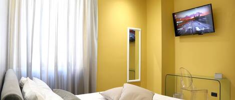 A hotel room with a bed, a television mounted on the wall, a mirror, and a glass partition.