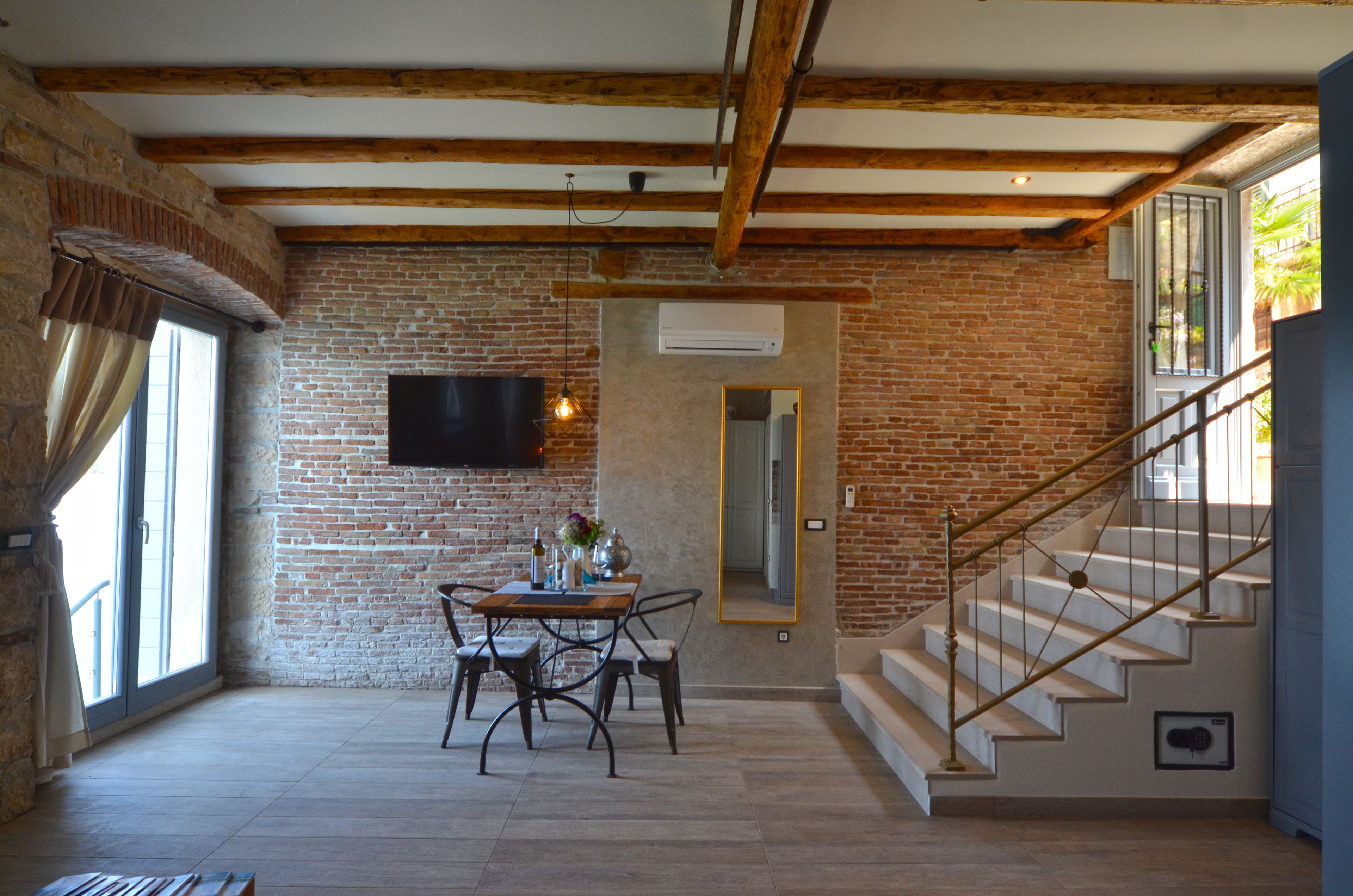 Une pièce spacieuse avec des poutres apparentes, un mur en briques, une table à manger avec des chaises, un téléviseur fixé au mur et un escalier menant à un niveau supérieur.