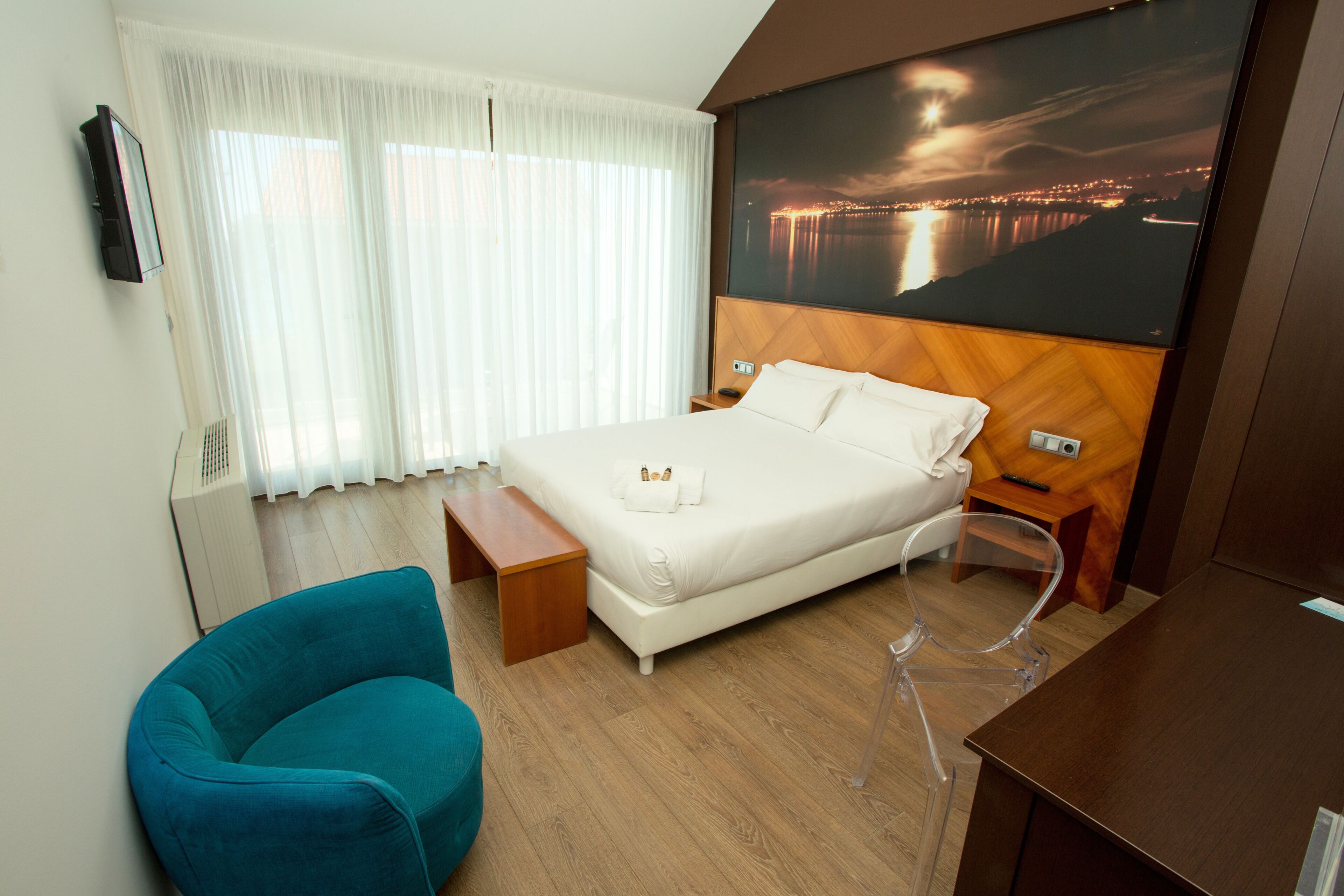 A hotel room with a bed, a chair, a desk, a large window with curtains, and a cityscape view on the wall.