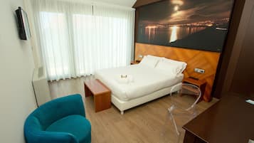 A hotel room with a bed, a chair, a desk, a large window with curtains, and a cityscape view on the wall.