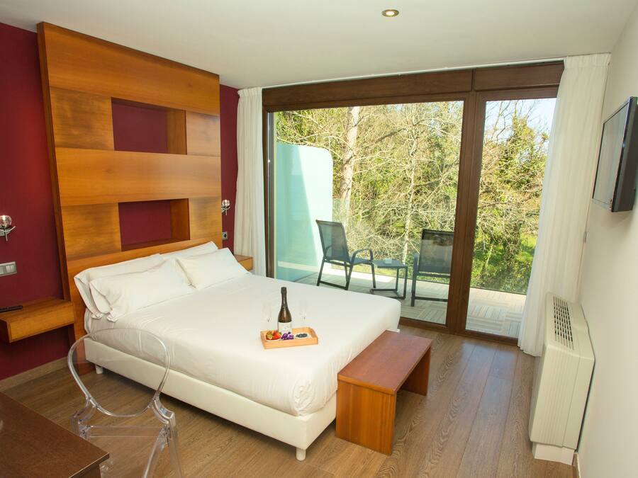 Habitación de hotel con una cama grande, una mesita de noche, un televisor, un balcón con una silla y un cabecero de madera.