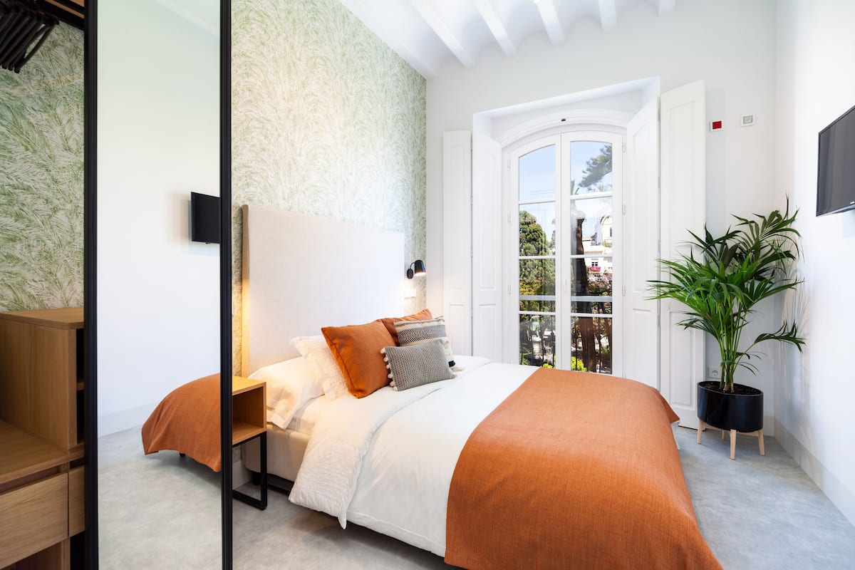 A bedroom with a bed, a nightstand, a potted plant, and a window with a view of the outside.