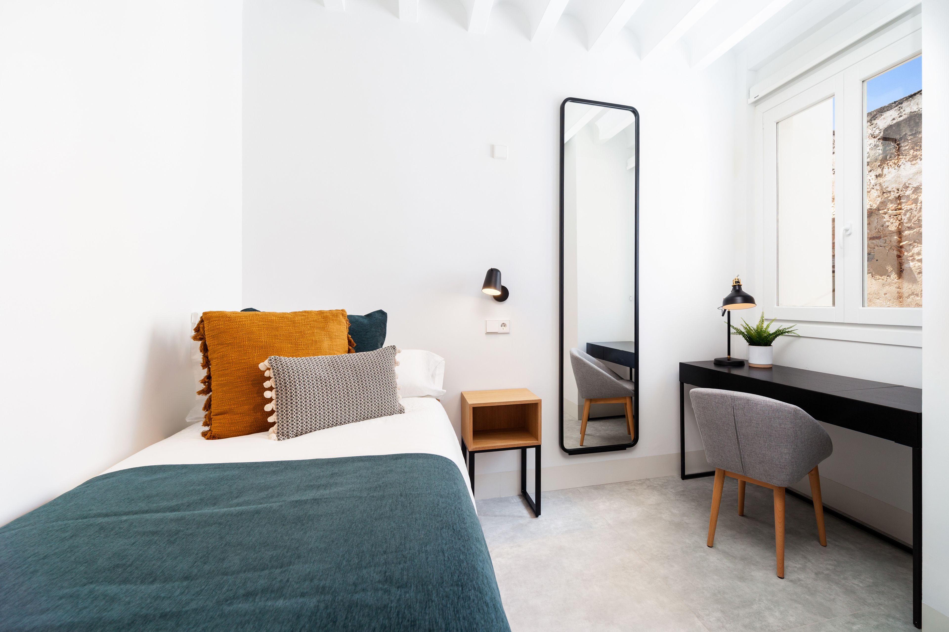 A modern bedroom with a bed, a desk, a chair, a mirror, and a window.