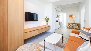 Uma sala de estar moderna com televisão de tela plana, armários de madeira, uma área de jantar e uma cozinha ao fundo.