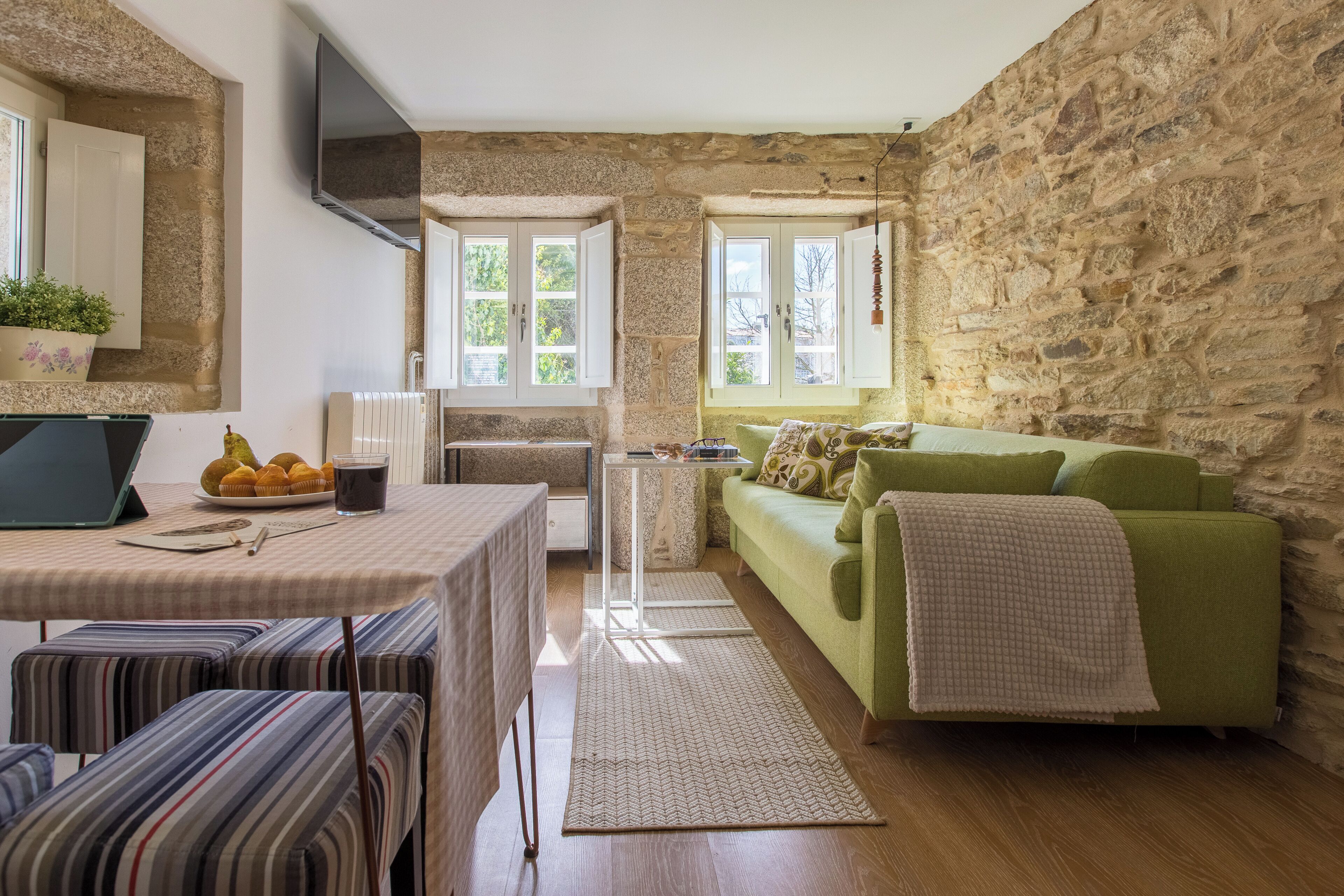 A cozy room with a stone wall, a green sofa, a striped bench, a table with a laptop, and a window with a view of trees.