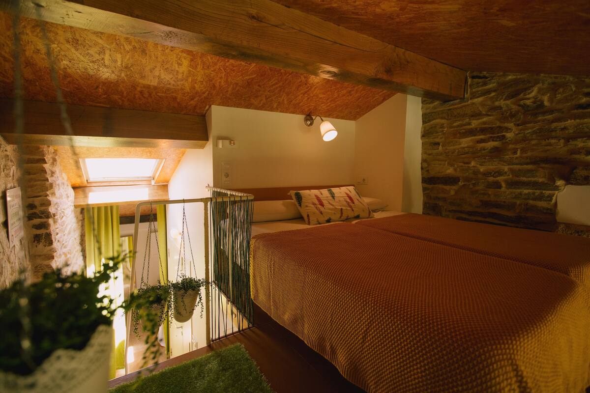Une chambre avec un lit, une lucarne, un mur en pierre et des plantes suspendues.