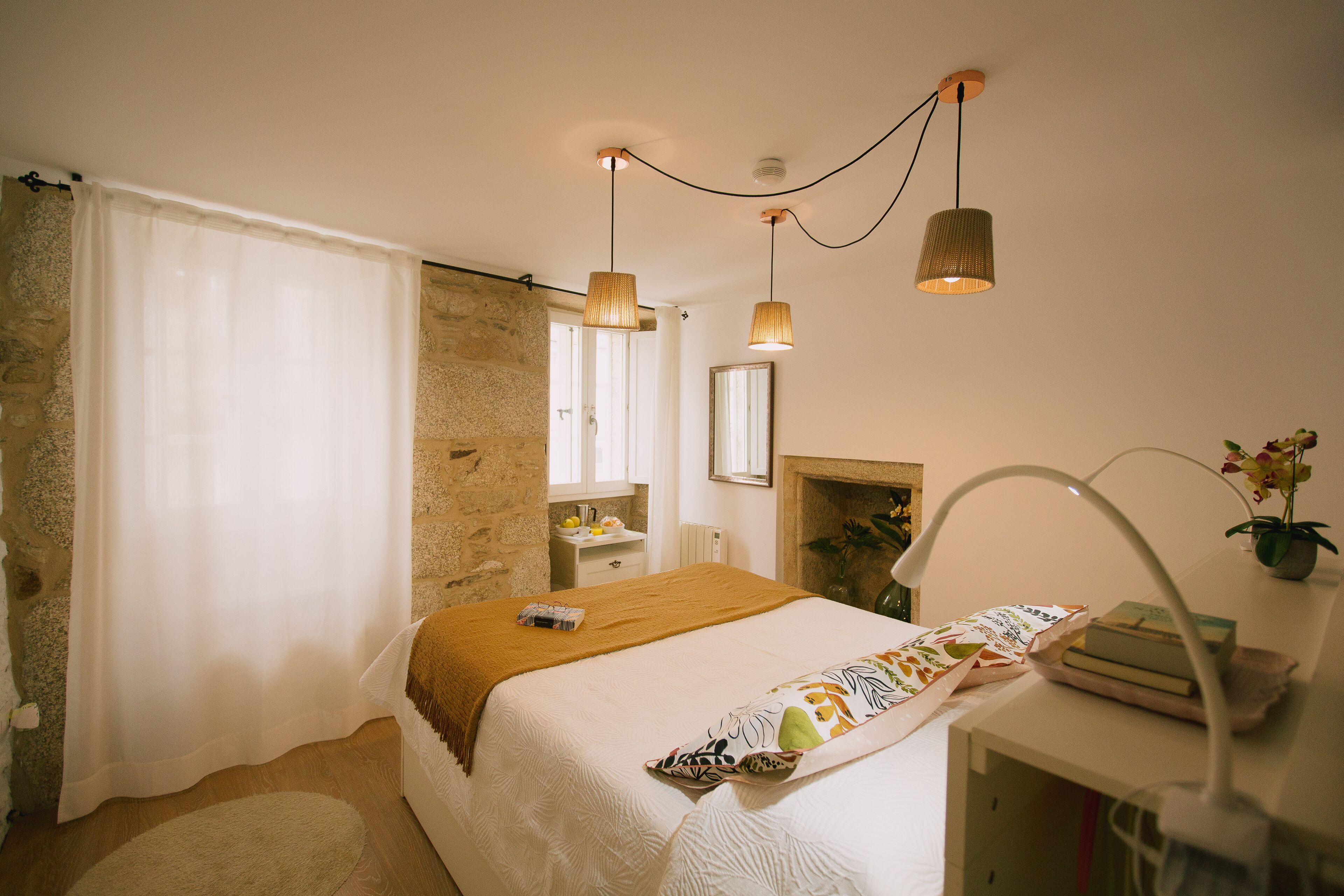 A bedroom with a bed, a bedside table, a mirror, a window with curtains, and a hanging light fixture.