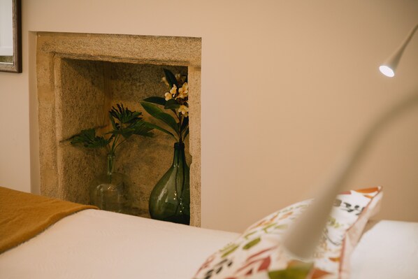 Una cama con sábana blanca y manta marrón, un jarrón verde con flores, y un cuadro colgado en la pared.