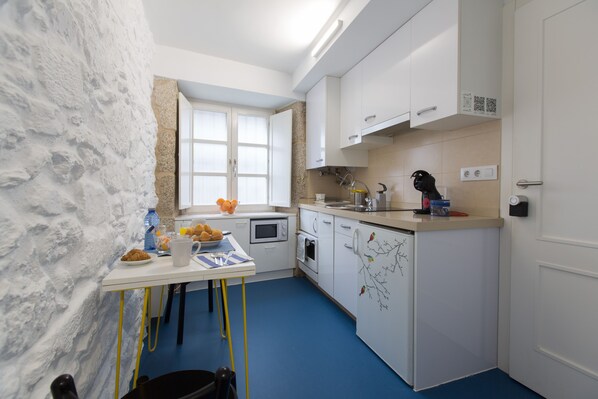 Cocina compacta con armarios blancos, microondas y refrigerador. Cuenta con una pequeña mesa de comedor con sillas, una ventana y piso azul.