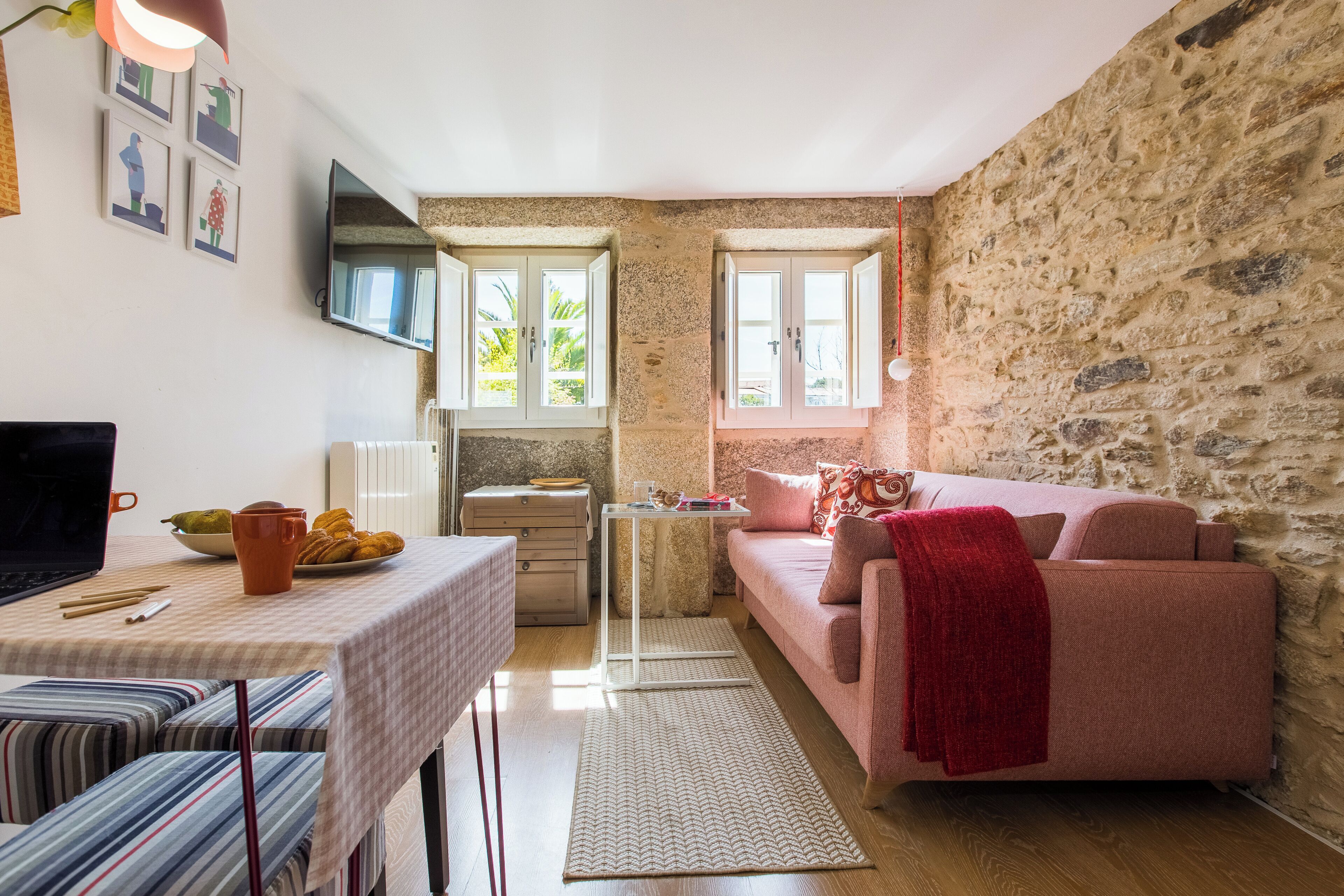 Una sala de estar acogedora con una pared de piedra, un sofá rosa, una mesa de comedor con una computadora portátil y un televisor montado en la pared.
